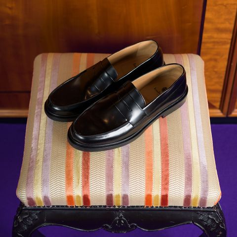 Une paire de mocassins noirs brillants repose sur un tabouret rembourré rayé dans les tons orange, violet et beige.