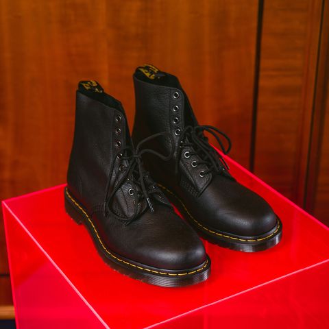 Une paire de bottes en cuir noir à lacets avec des surpiqûres décoratives jaunes repose sur un présentoir rouge brillant devant un fond en bois.