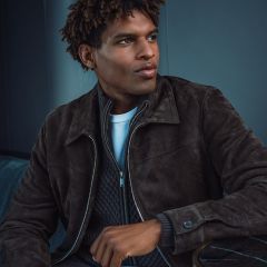 Un homme portant une veste en daim marron foncé, un pull gris et une chemise blanche est assis dans une pièce, regardant sur le côté avec une expression pensive.