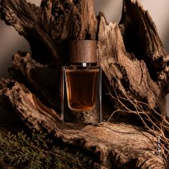 Un flacon de parfum en verre avec un bouchon en bois est posé au milieu d'écorce d'arbre brute et de branches séchées, dégageant ainsi une atmosphère rustique et naturelle.