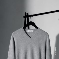 Un pull gris clair à col en V est suspendu à un cintre noir devant un fond uni, en partie dans l'ombre, en partie dans la lumière.