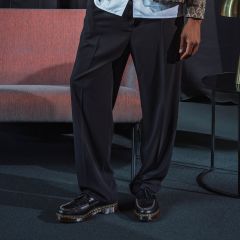 Une personne se tient à l'intérieur et porte un pantalon noir à jambe large et des chaussures compensées noires ; à côté d'elle se trouvent un canapé rose et une petite table ronde.