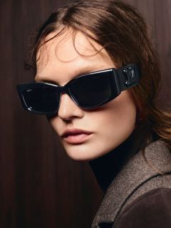 Une femme aux longs cheveux bruns, portant de grandes lunettes de soleil noires et un manteau marron, posant devant un fond en bois sombre.