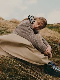 Une personne vêtue d'un manteau à carreaux et d'une longue jupe est assise dehors dans les hautes herbes, regardant pensivement la caméra.