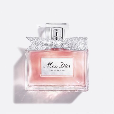 Un flacon en verre transparent de « Miss Dior Eau de Parfum », rempli d'un liquide rose et orné d'un nœud argenté à motifs autour du col du flacon, sur un fond blanc uni.