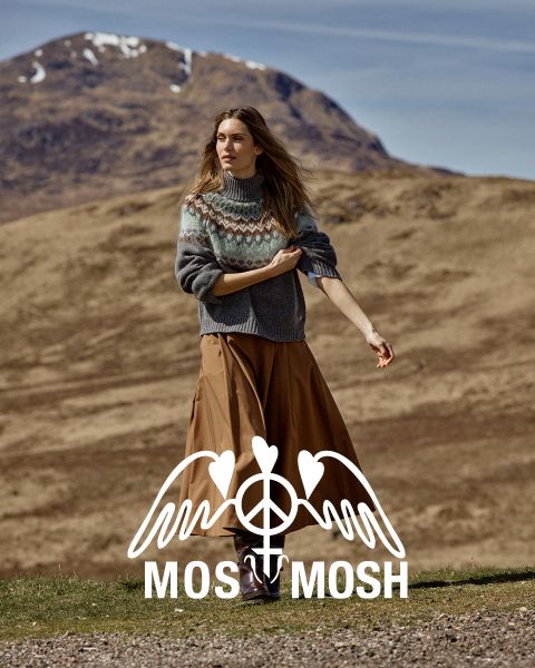 Une femme vêtue d'un pull à motifs et d'une jupe marron se tient à l'extérieur, avec des collines et une montagne derrière elle. Le logo "MOSS MOSH" est visible en bas de l'image.