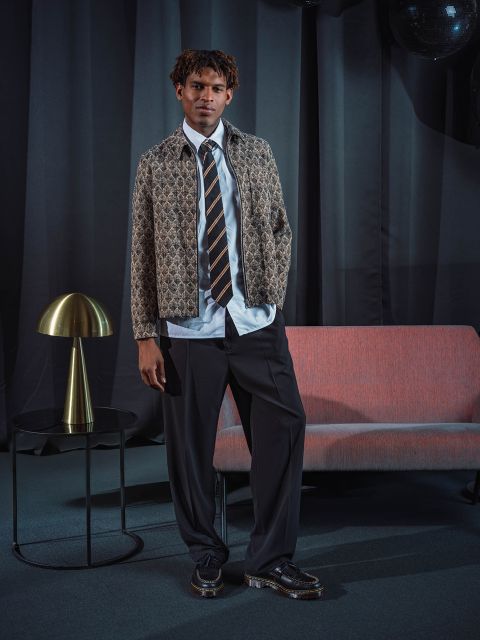 Un homme en veste à motifs, cravate et pantalon de costume se tient à côté d'un canapé rose et d'une lampe dorée devant des rideaux sombres.