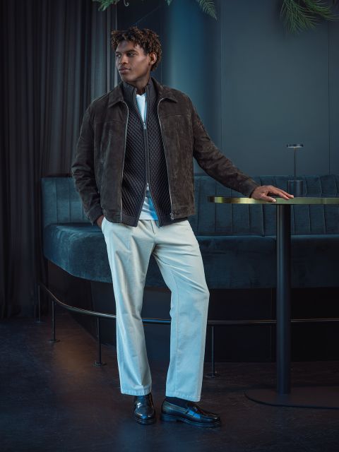 Un homme dans une veste marron et un pantalon blanc se tient à côté d'une table ronde dans un salon moderne, faiblement éclairé, avec des sièges et des rideaux bleu foncé.