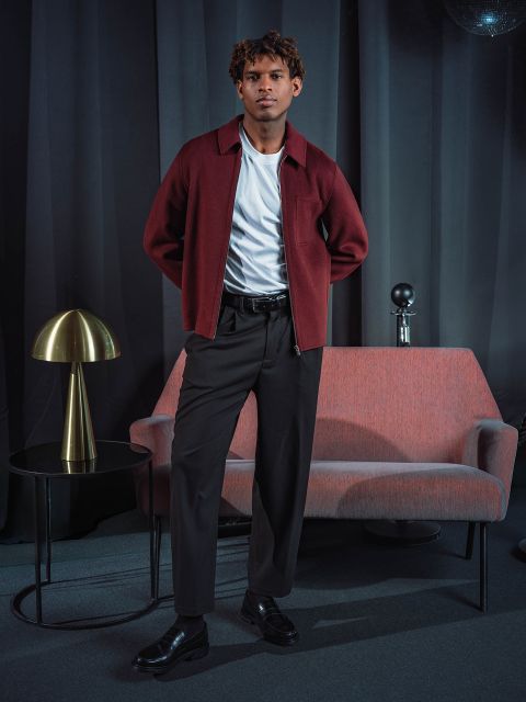 Un homme vêtu d'une veste bordeaux et d'un pantalon noir se tient à côté d'un canapé rose, sur lequel se trouve une table ronde avec une lampe dorée, devant des rideaux sombres.