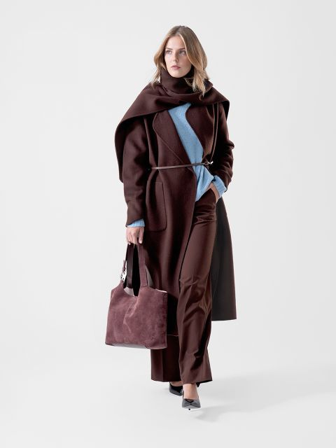 Une femme dans un manteau marron foncé et un pantalon marron foncé, ainsi qu'un pull bleu, tenant un sac marron assorti à la main et marchant avec assurance devant un simple fond blanc.