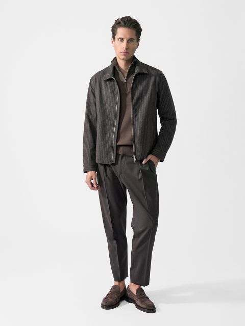 Un homme, portant une veste rayée foncée, un pull marron, un pantalon foncé et des mocassins marron, se tient devant un fond blanc uni.