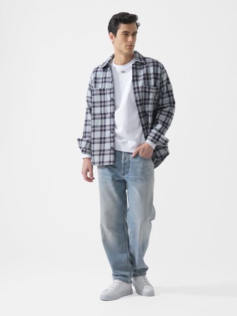 Un homme, portant un t-shirt blanc, une chemise à carreaux, un jean bleu clair et des baskets blanches, se tient devant un simple fond blanc.
