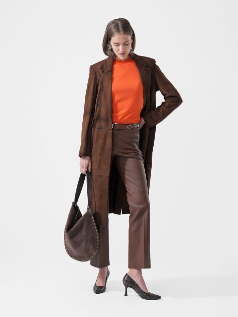 Une femme dans un manteau marron, un haut orange, un pantalon marron et des escarpins tient un sac à main marron et se tient devant un fond blanc uni.