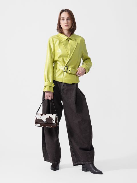 Une femme est debout, vêtue d'une veste vert citron ceinturée, d'un jean large bleu foncé et de bottes noires, et tenant un petit sac à main à motif vache.