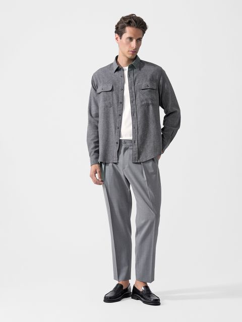Un homme, portant une chemise grise boutonnée, un t-shirt blanc, un pantalon de costume gris et des mocassins noirs, et se tenant devant un simple fond blanc.