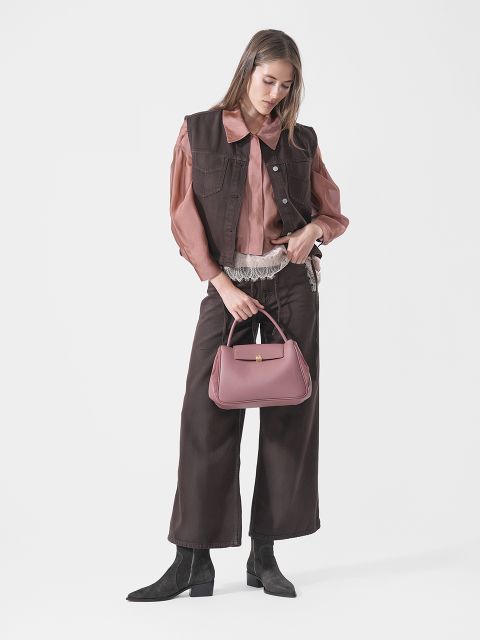 Une femme en gilet et pantalon marron, blouse rose et bottes noires tient un sac à main rose et se tient devant un fond blanc uni.