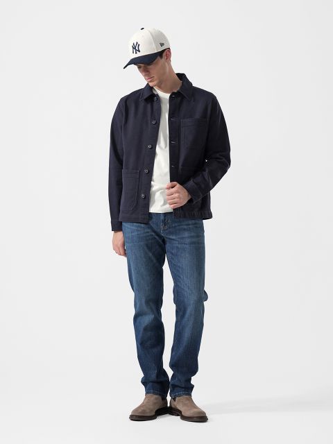 Un homme portant une veste bleu marine, un t-shirt blanc, un jean bleu, des bottes grises et une casquette de baseball NY blanche, se tient devant un fond blanc uni.