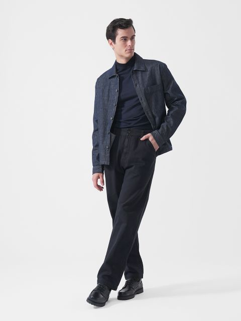 Un homme portant une veste en jean foncée, un col roulé bleu marine, un pantalon noir et des chaussures noires, se tient devant un fond blanc uni et regarde vers la gauche.