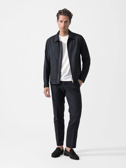 Devant un fond blanc se tient un homme portant une veste foncée à rayures, un pantalon assorti, un t-shirt blanc et des mocassins noirs.