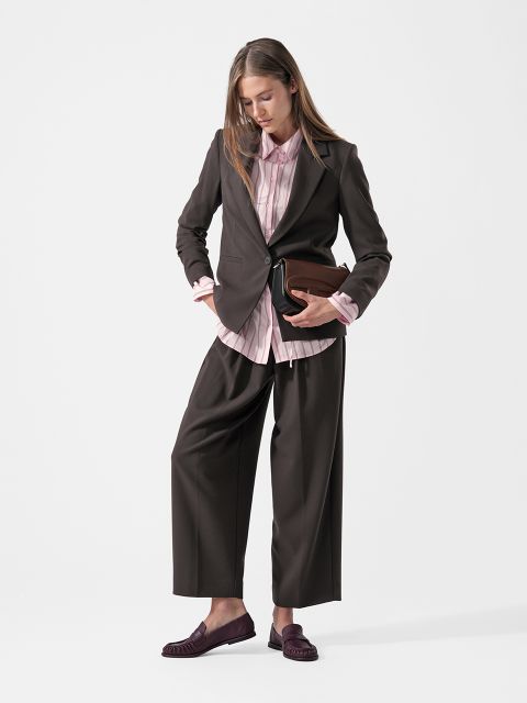 Une femme dans un tailleur-pantalon sombre, une chemise rose et des mocassins tient une pochette marron à la main, se tient devant un fond blanc uni et regarde vers le bas.