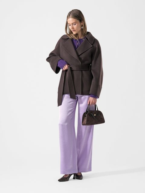 Une femme dans un manteau marron et un pantalon violet tient un petit sac à main marron et regarde vers le bas. Elle porte des chaussures noires, et sous son manteau, des manches violettes dépassent.