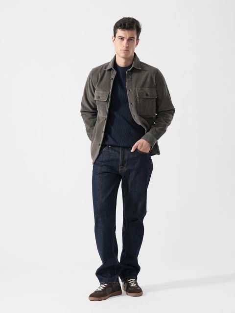 Un homme dans une veste grise, un jean bleu foncé et des baskets marron se tient devant un fond blanc uni et a les mains dans les poches.
