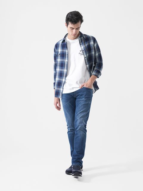 Un homme portant une chemise à carreaux bleue, un t-shirt blanc, un jean bleu et des baskets, marchant devant un simple fond blanc et regardant vers le bas.