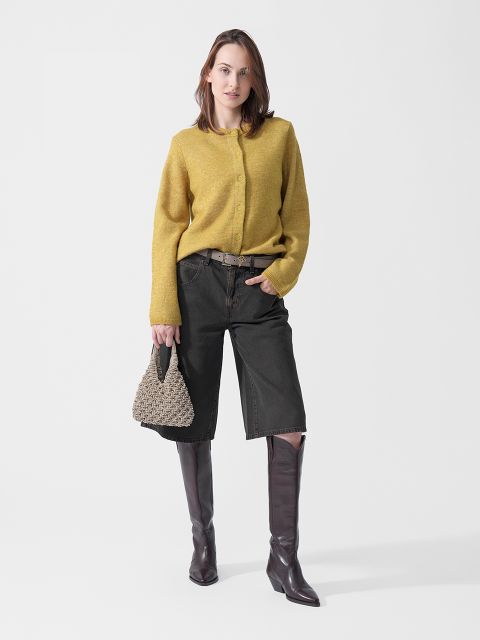 Une femme portant un pull jaune, un short noir arrivant aux genoux et des bottes marron, et tenant un sac à main tressé, se tient devant un fond blanc.
