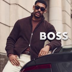 Un homme en veste marron, avec des lunettes de soleil et un pantalon clair, est appuyé contre une voiture à côté de l'inscription « BOSS ».