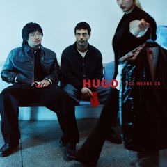 Deux hommes sont assis sur un banc, tandis qu'une personne au premier plan tient un sac à vêtements avec l'inscription « HUGO RED MEANS GO ».