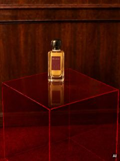 Sur une table rouge transparente devant un mur en bois et un tapis rouge se trouve une bouteille de parfum Bel-Ami.