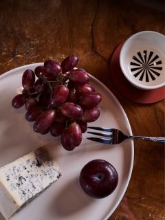Une assiette de raisins rouges, une tranche de fromage bleu et une prune ; on peut également voir une fourchette et une tasse sur une table en bois.
