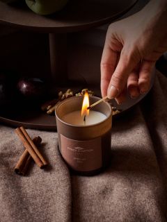 Une main allume une bougie marron "African Tea" de Finch Hatton, posée à côté de bâtons de cannelle, de prunes et de noix sur un tissu doux et marron.