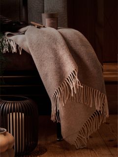 Une couverture beige à franges drapée sur une chaise, à côté une bougie dans une lanterne noire et une tasse de café sur le parquet en bois.