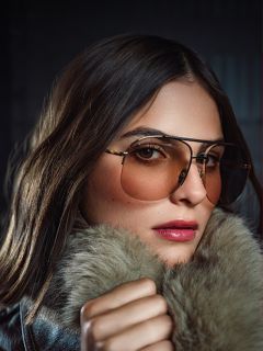 Une femme aux longs cheveux bruns porte des lunettes d'aviateur teintées et un manteau à col de fourrure, regardant la caméra avec confiance sur un fond sombre.