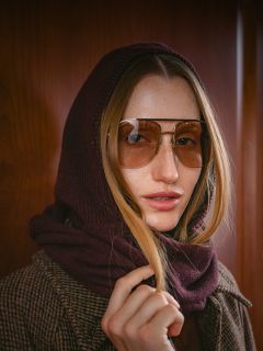 Une femme aux cheveux longs, portant des lunettes d'aviateur, un foulard marron sur la tête et un manteau à carreaux marron, se tient devant un mur en bois.