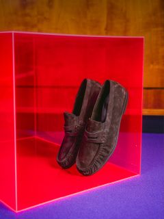 Mocassins en daim marron, présentés dans un cube rose transparent, sur un fond en bois et sur une base violette.
