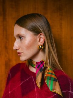 Femme en pull rouge avec une écharpe à carreaux colorée et des boucles d'oreilles dorées, regardant vers la gauche, devant un fond en bois.