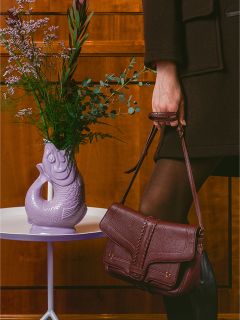 Une personne vêtue de vêtements sombres tient un sac à main bordeaux à côté d'un vase violet en forme de poisson avec des fleurs sur une table blanche.
