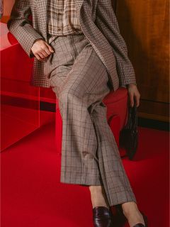 Une personne en costume à carreaux est assise les jambes croisées sur une chaise rouge, tenant un sac sombre, sur un tapis rouge.
