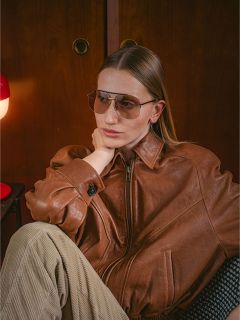 Une femme en veste de cuir marron et lunettes de soleil est assise à l'intérieur, le menton appuyé sur la main, à côté d'une lampe rouge et d'une armoire en bois.