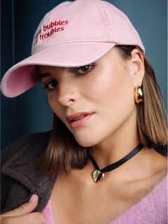 Une femme aux cheveux bruns lisses porte un bonnet rose, des boucles d'oreilles dorées, un collier en forme de cœur et un haut rose et moelleux, et regarde la caméra devant un fond bleu.