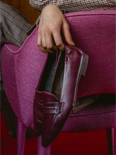 Une personne en pantalon à carreaux est assise sur une chaise rose à surface texturée, tenant un mocassin bordeaux.