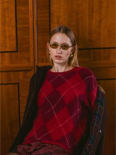 Une femme portant un pull argyle rouge, des lunettes de soleil et des boucles d'oreilles dorées est assise devant un mur lambrissé et regarde l'appareil photo.