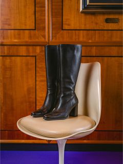 Une paire de bottes en cuir noires, hauteur genou, à talons blocs, est posée sur une chaise moderne de couleur crème devant un mur lambrissé de bois.