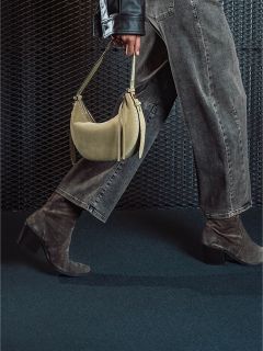 Une personne en jean gris et bottes à talons en daim tient un sac à main beige et passe devant un fond sombre texturé.