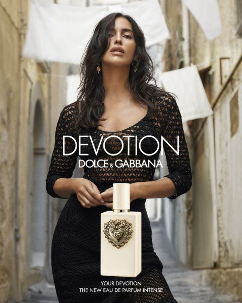 Une femme vêtue de noir se tient dans une ruelle étroite derrière une bouteille de parfum « Dolce & Gabbana Devotion ». Du linge blanc est suspendu en arrière-plan.