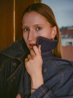 Une femme aux cheveux lisses porte une veste en jean foncée dont elle a relevé le col, et regarde l'appareil photo avec un visage inexpressif.