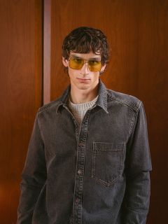 Un homme aux cheveux ondulés, portant des lunettes de soleil jaunes, une chemise en jean grise et un pull clair, se tient devant un mur en bois et regarde l'appareil photo.