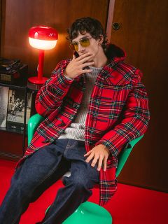 Un homme en manteau à carreaux rouge et lunettes de soleil est assis sur une chaise verte à côté d'une lampe rouge et tient sa main près de son visage dans une pose élégante.
